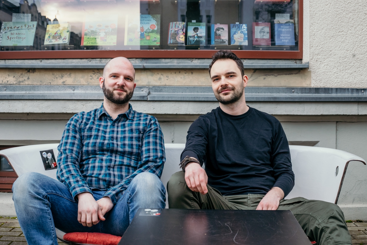 Alexander Senf und Alexander Büttner sitzen vor dem Buchladen und lächeln in die Kamera.